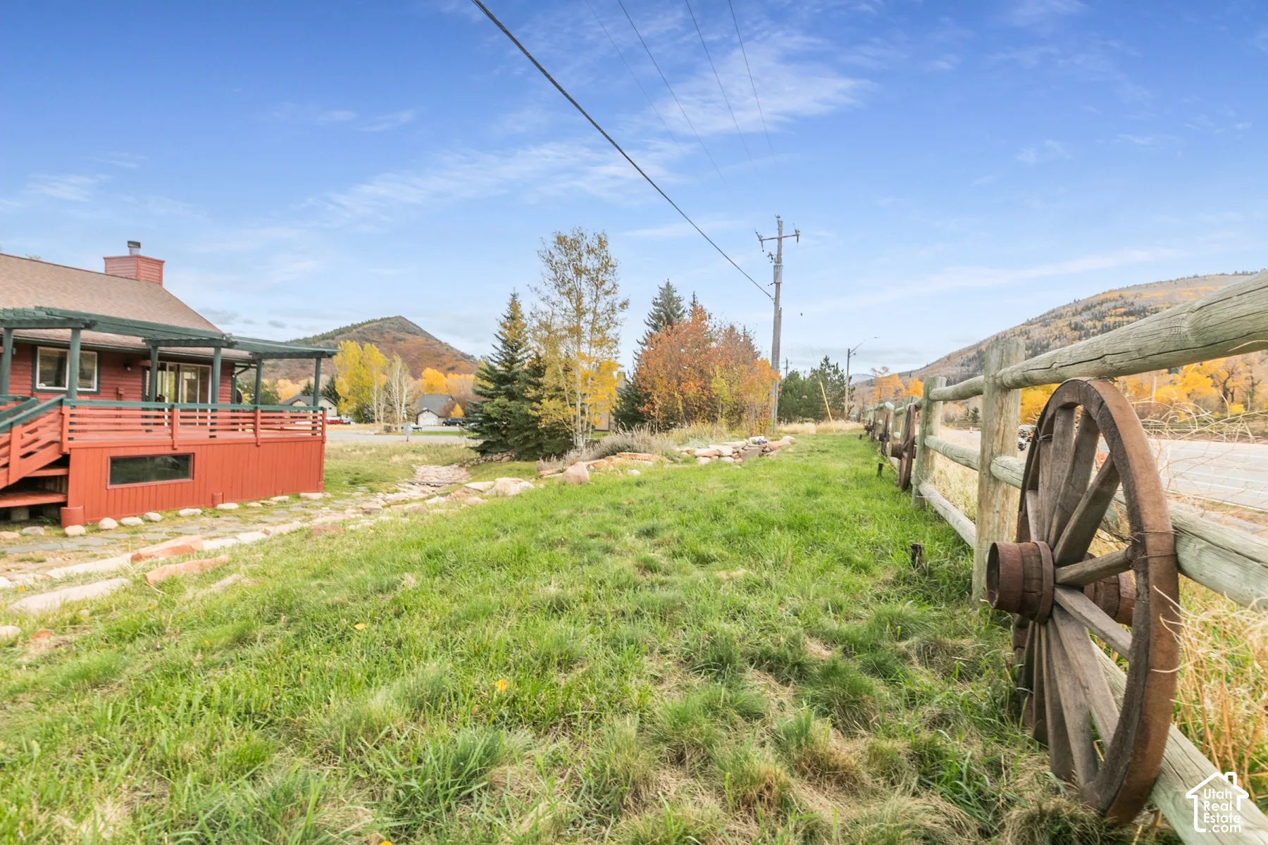 Exterior grounds featuring a red-toned wooden deck with a covered pergola, a rustic split-rail fence adorned with antique wheel accents, expansive lawn, and mountain topography