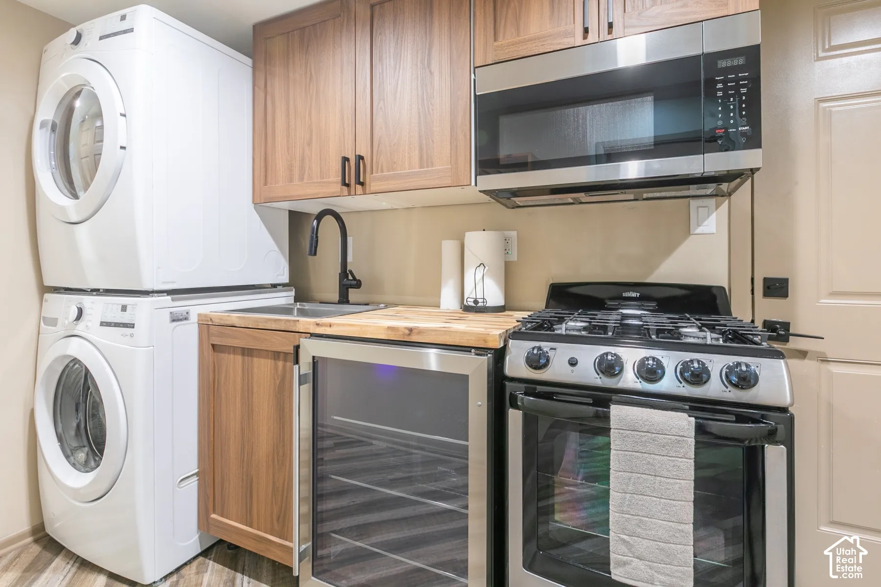 Stacked washer and dryer, wood-finish cabinetry, stainless steel gas range, built-in beverage refrigerator, and wood-block countertop with integrated sink