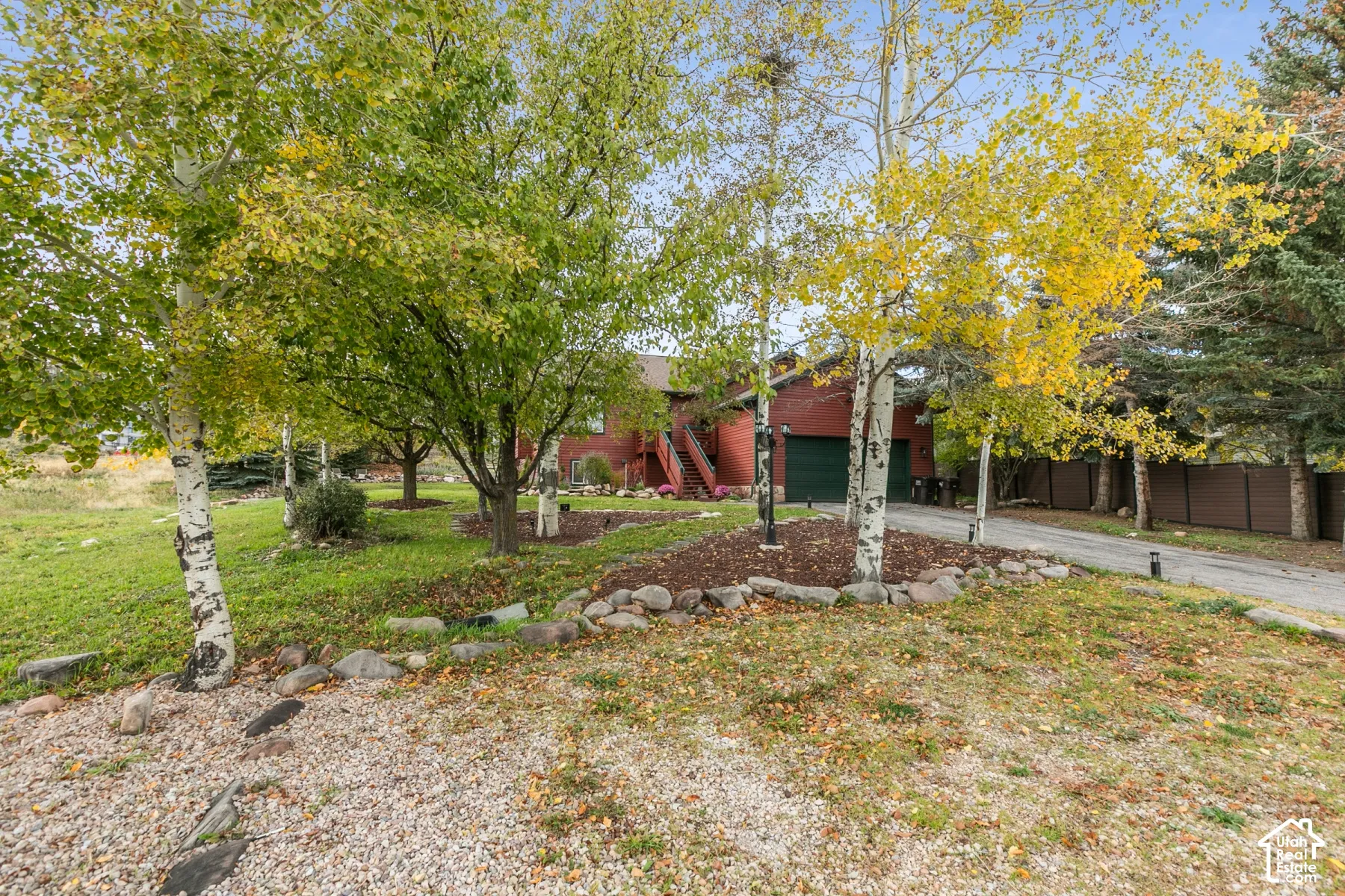 Red wood-siding exterior featuring a two-car garage, mature landscaping, river rock borders, and a paved driveway