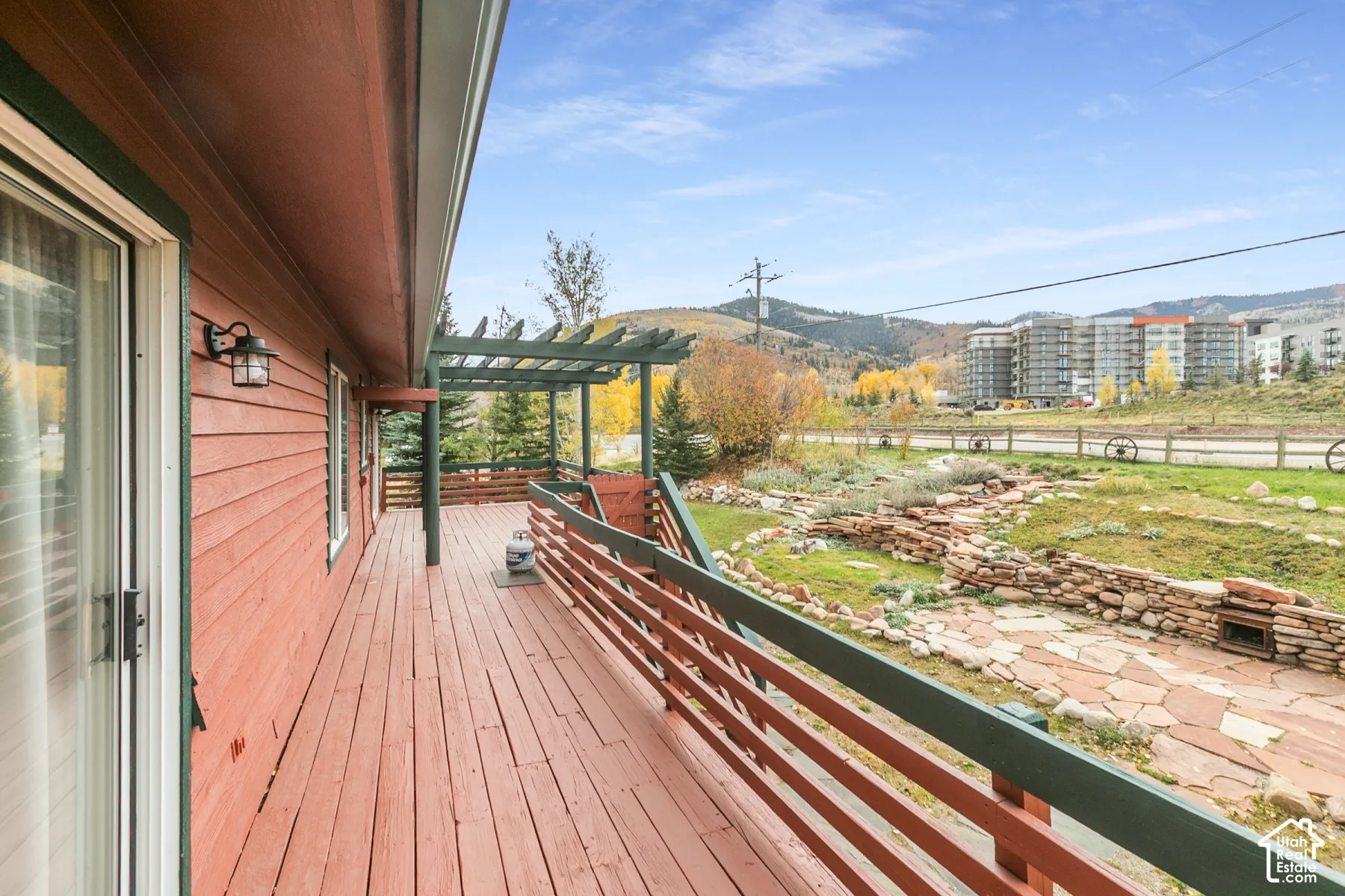 Extended exterior deck with wood-finish decking, pergola structure, horizontal slat railing, wall-mounted sconce lighting, and natural stone hardscaping