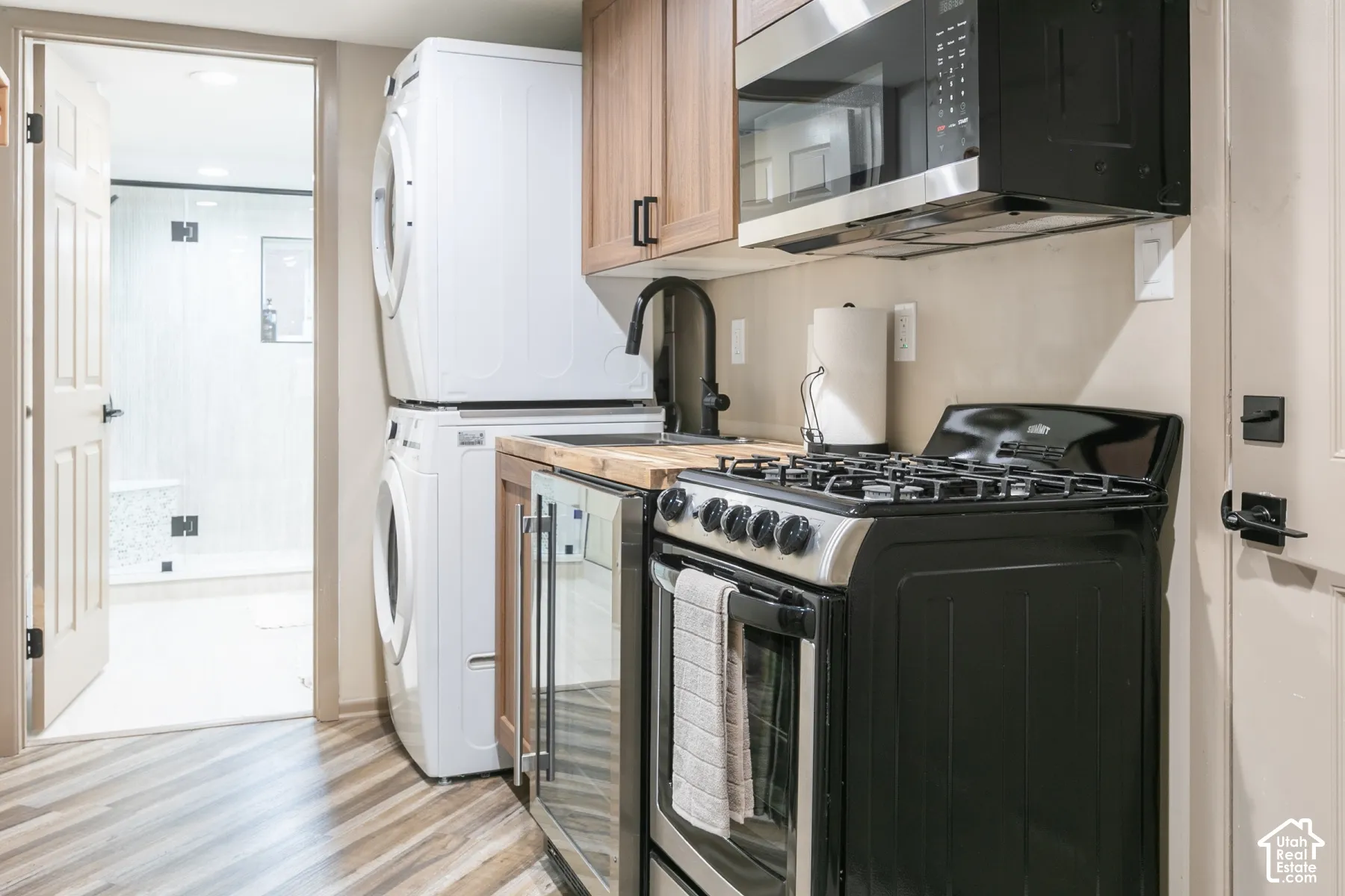 Kitchenette featuring wood-finish cabinetry, black gas range, built-in microwave, and wood-patterned flooring, with laundry area containing a stacked washer and dryer unit