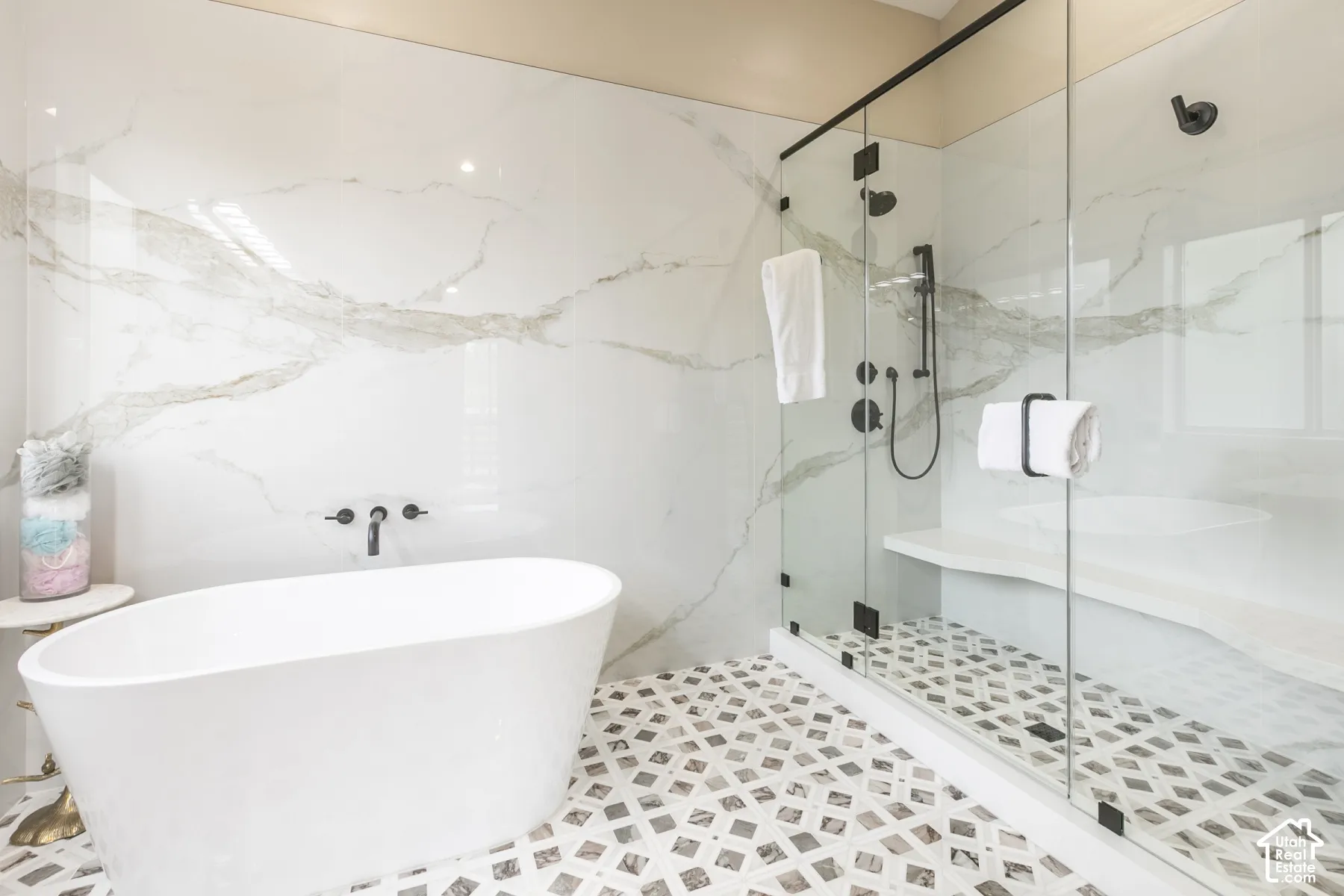 Freestanding soaking tub with wall-mounted matte black faucet, large-format stone-finish wall tile, frameless glass-enclosed shower with built-in bench, and geometric patterned floor tile
