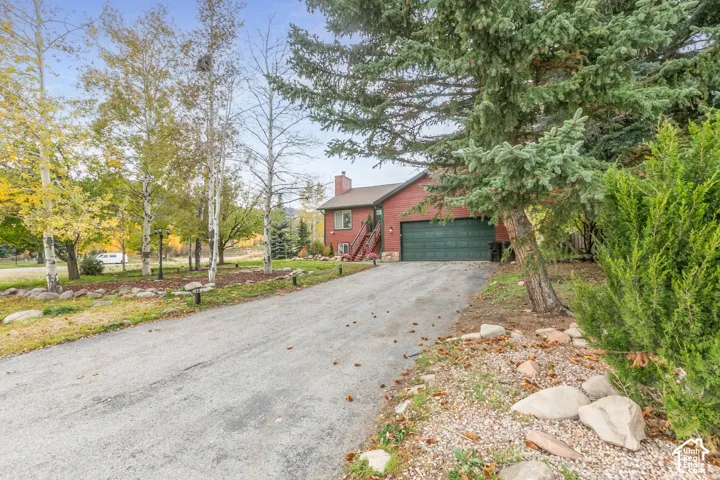 Red-sided exterior with stone foundation detailing, asphalt-paved driveway, two-car garage, and mature evergreen and aspen landscaping