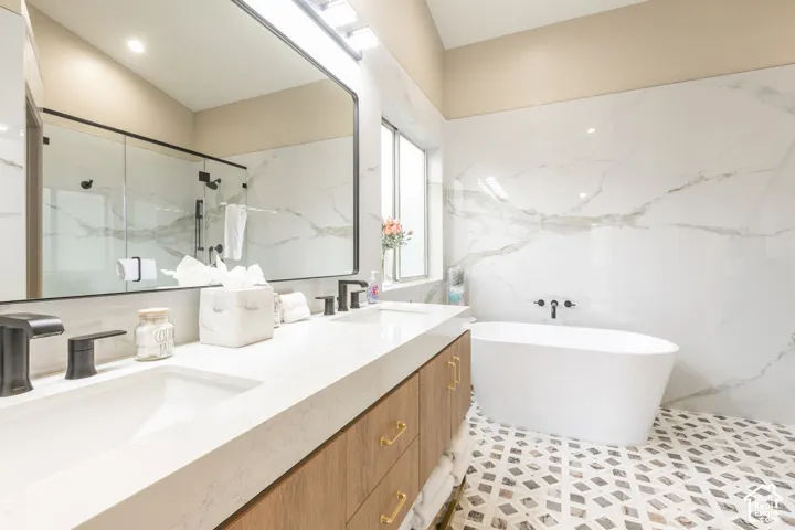 Dual-sink vanity with a solid-surface countertop and matte black fixtures, freestanding soaking tub, expansive wall-to-wall tiling, geometric patterned stone-finish floor, and large-format framed mirror