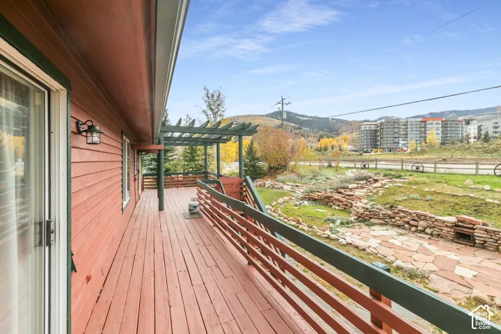 Extended exterior deck with wood-finish decking, pergola structure, horizontal slat railing, wall-mounted sconce lighting, and natural stone hardscaping
