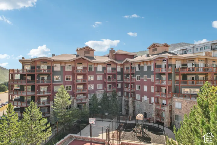 Multi-story building exterior featuring vertical siding, stone masonry accents, multiple wooden balconies, and mountain surroundings, including a fenced court and playground area