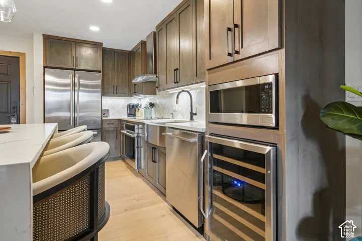 Kitchen featuring wood-finish cabinetry, stainless steel appliances, a farmhouse-style sink with a matte black faucet, built-in beverage refrigerator, and light-toned wood-finish flooring
