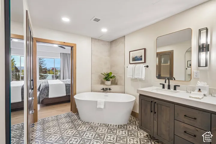 Freestanding soaking tub with wall-mounted matte black faucet, custom dark wood cabinetry with quartz countertop, geometric patterned tile flooring, and rectangular mirror with vertical integrated wall sconce