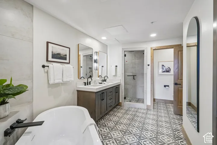 Freestanding soaking tub with matte black wall-mounted faucet, dual-sink vanity with stone countertop, glass-enclosed shower, and patterned tile flooring
