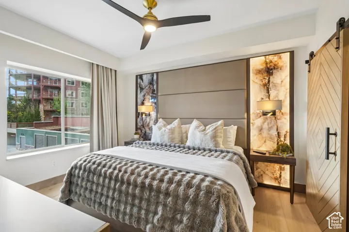 Custom upholstered headboard wall featuring integrated backlighting, stone accent panels, and chevron-patterned wooden sliding door with matte black hardware, complemented by wood-finish flooring and a contemporary multi-blade ceiling fan