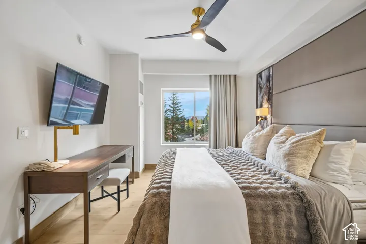 Light-toned wood-finish flooring, ceiling fan with integrated lighting, window with mountain backdrop, and floor-to-ceiling padded headboard wall paneling