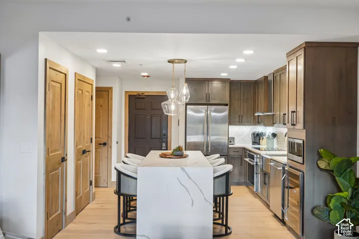 Kitchen featuring an island with a stone-finish waterfall countertop, stainless steel appliances, stained wood cabinetry, recessed lighting, and pendant fixtures
