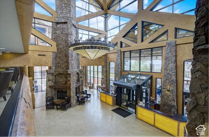 Vaulted timber-frame ceiling featuring floor-to-ceiling glass windows, prominent stone-clad columns, a central stone-surround fireplace, and a large custom metal chandelier