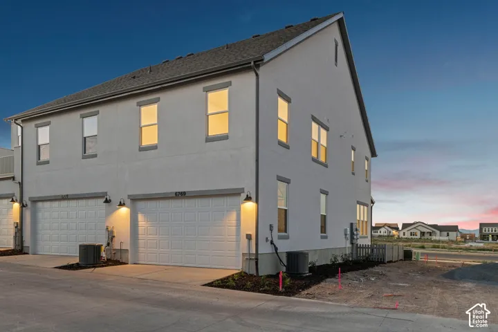 Multi-level exterior featuring light-toned stucco siding, attached two-car garage, asphalt shingle roof, and wall-mounted exterior lighting fixtures