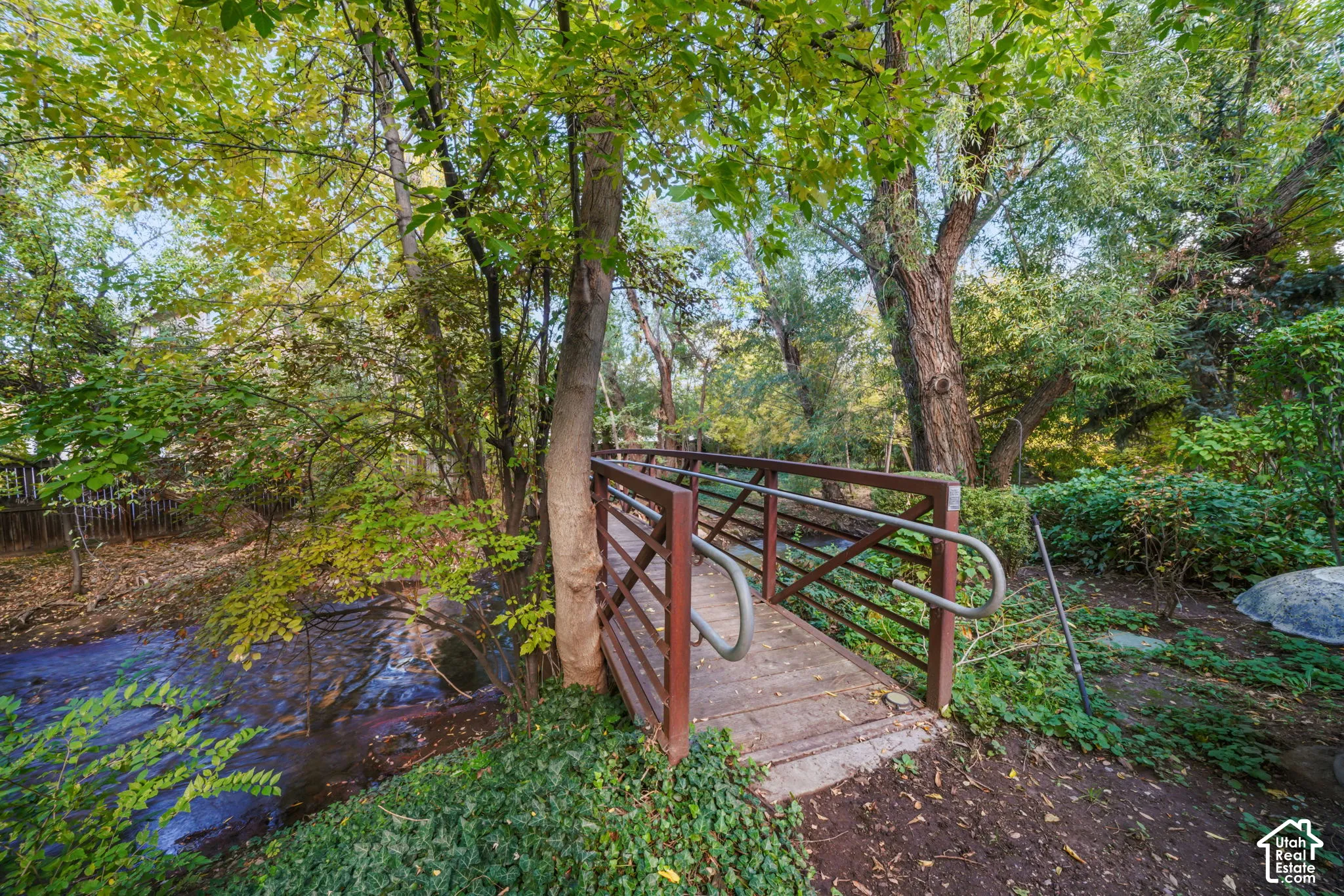 Pedestrian bridge featuring a wooden deck, metal framing, and railings spanning a natural water feature surrounded by mature trees and vegetation