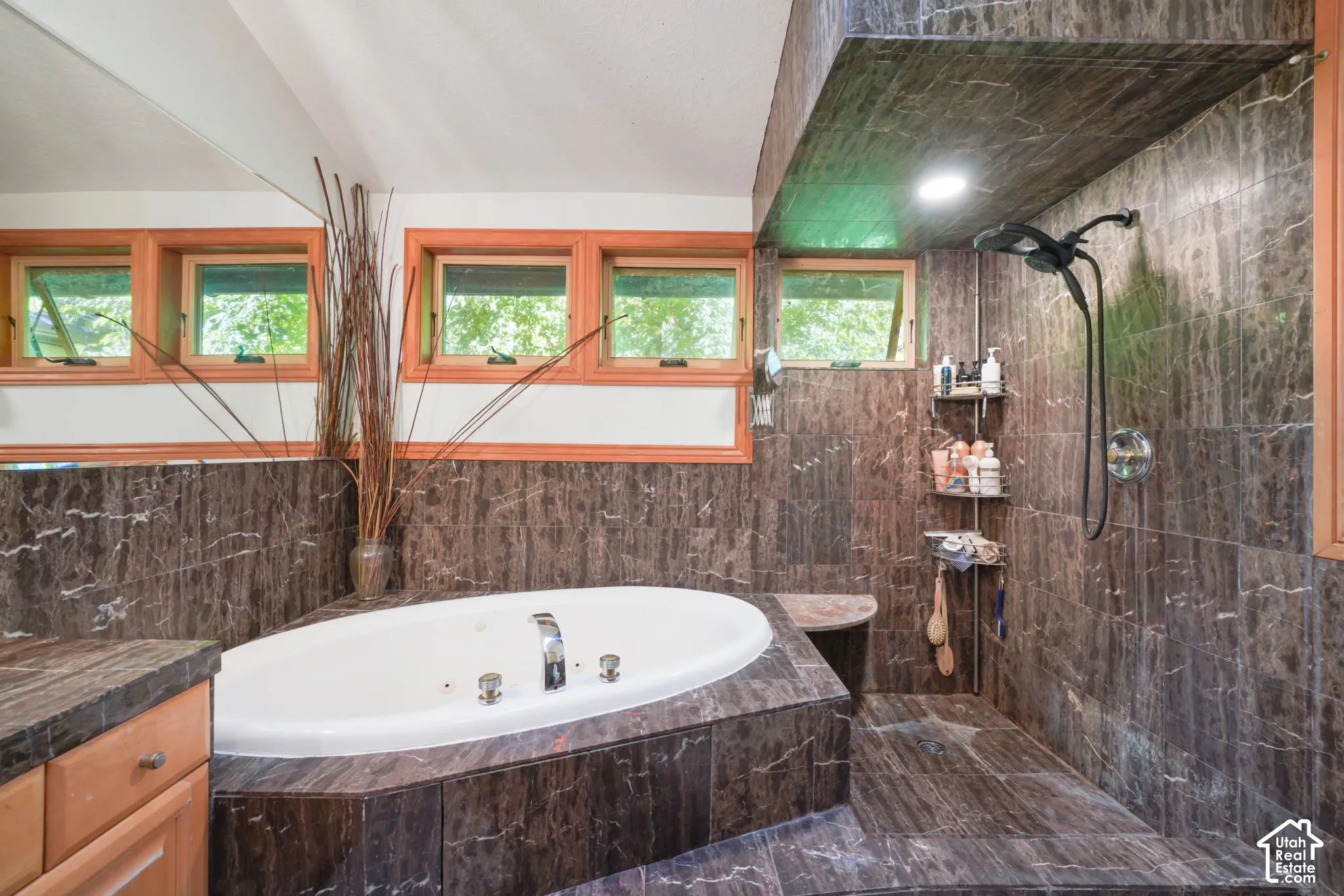 Bathroom featuring a jetted oval soaking tub, natural stone tile surround, walk-in shower with integrated bench seating, and wood-framed awning windows