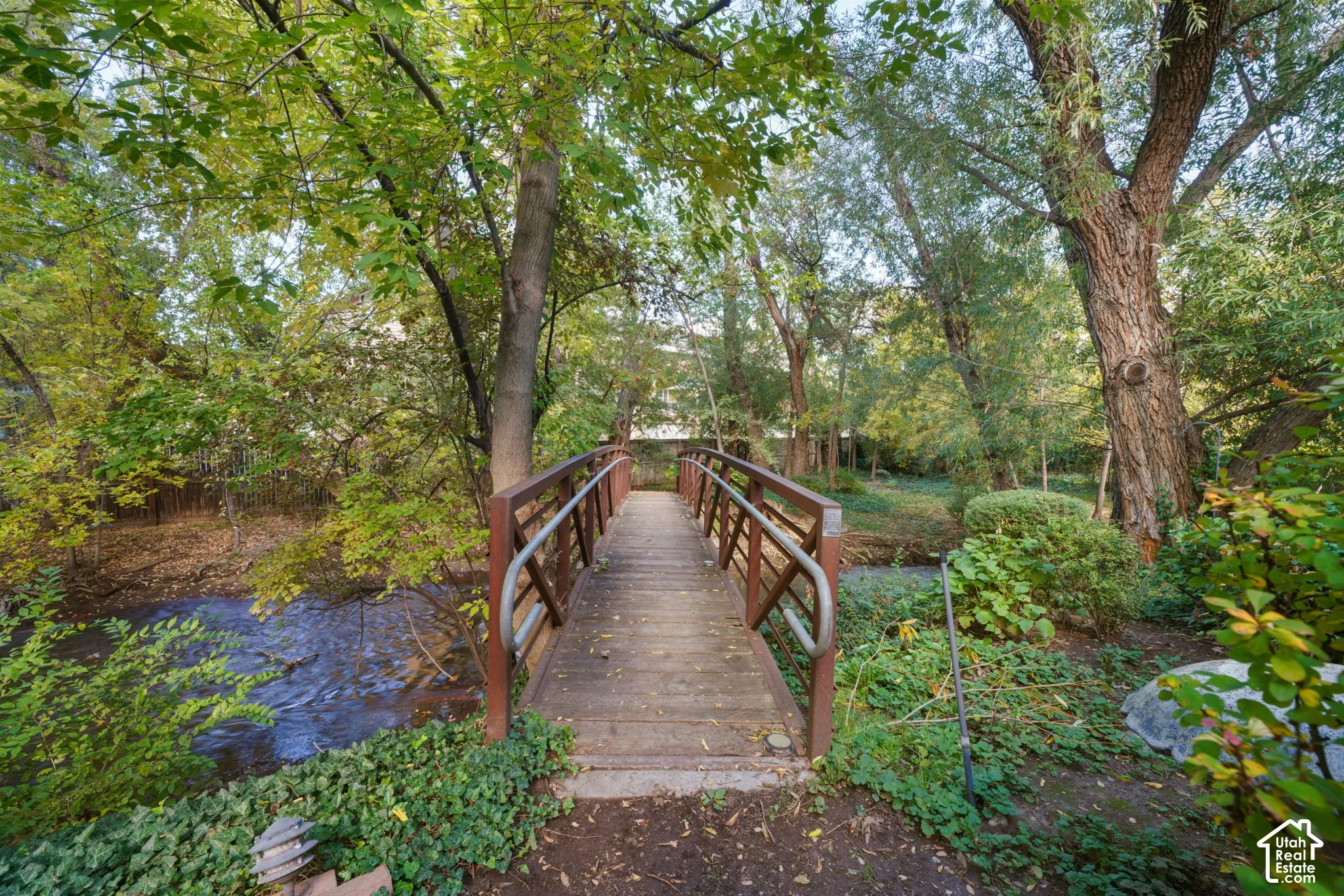 Pedestrian bridge with wooden decking and metal handrails spanning a natural water feature, complemented by mature trees and established landscaping