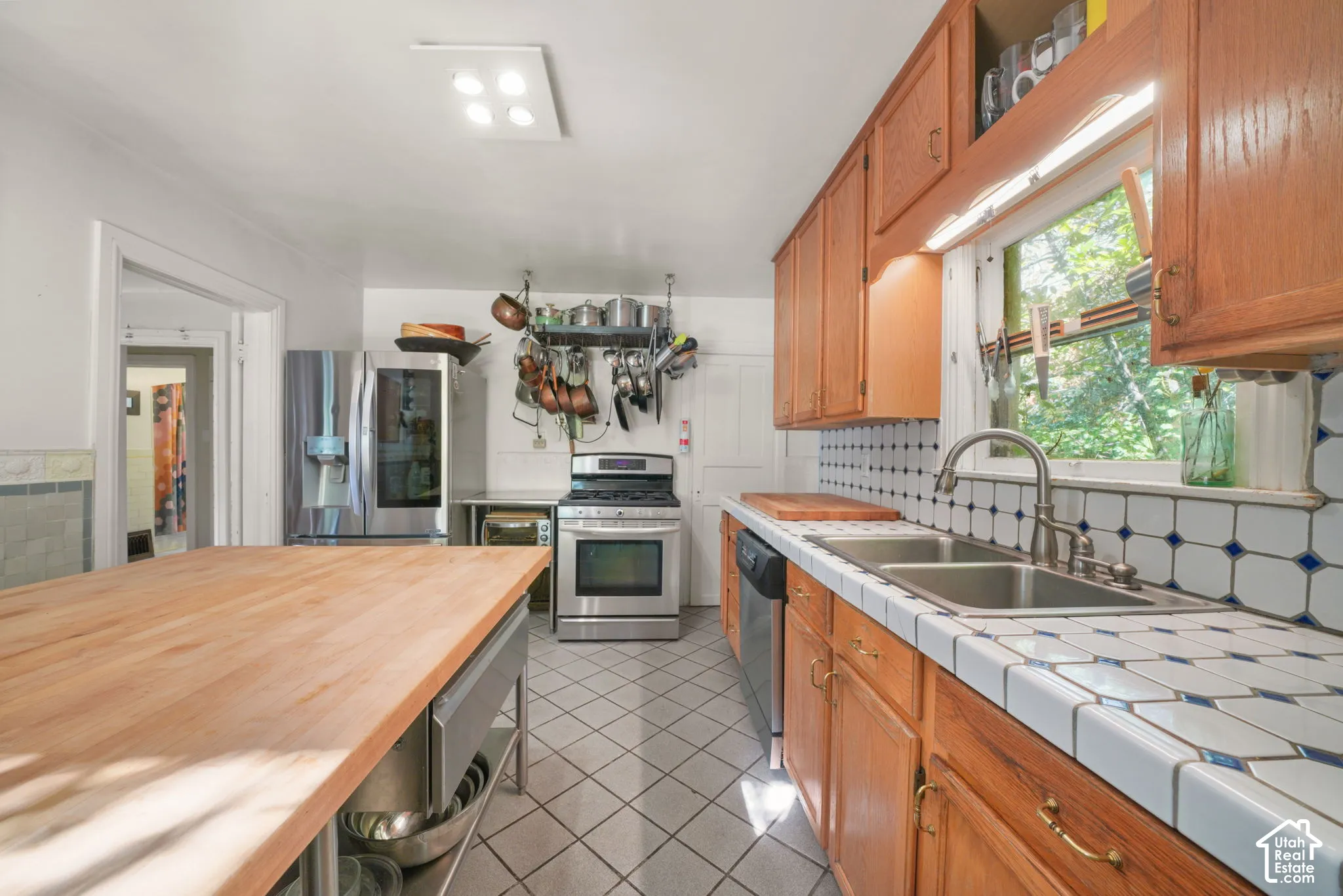 Kitchen featuring wood cabinetry, butcher block island countertop, tiled countertops with decorative accents, stainless steel appliances, and tiled flooring