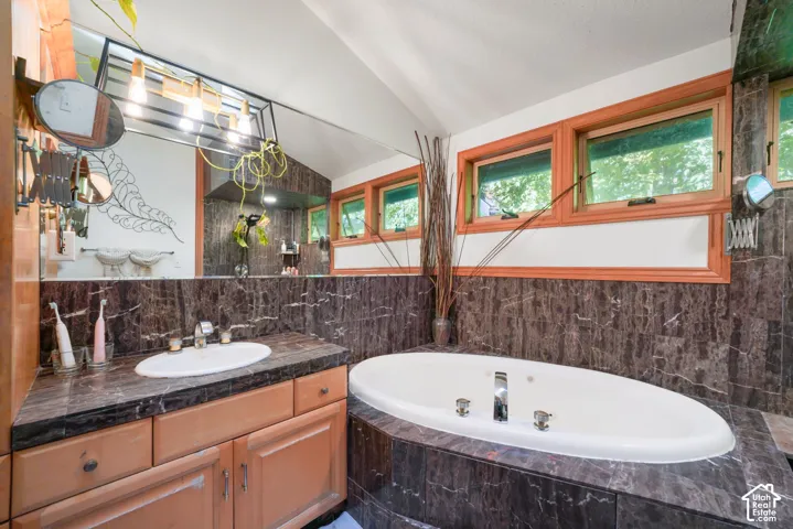 Bathroom featuring dark stone tile surround, oval jetted soaking tub, wood cabinetry, stone-topped vanity with undermount sink, and wood-framed windows