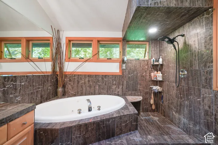 Bathroom featuring a jetted oval soaking tub, natural stone tile surround, walk-in shower with integrated bench seating, and wood-framed awning windows