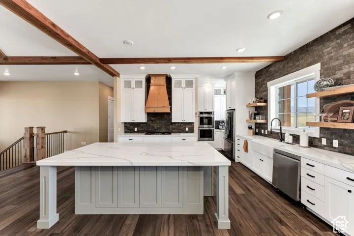 Kitchen featuring a large central island with quartz countertops, custom white cabinetry, exposed wood ceiling beams, a decorative wood range hood, and dark stone-finish wall accents