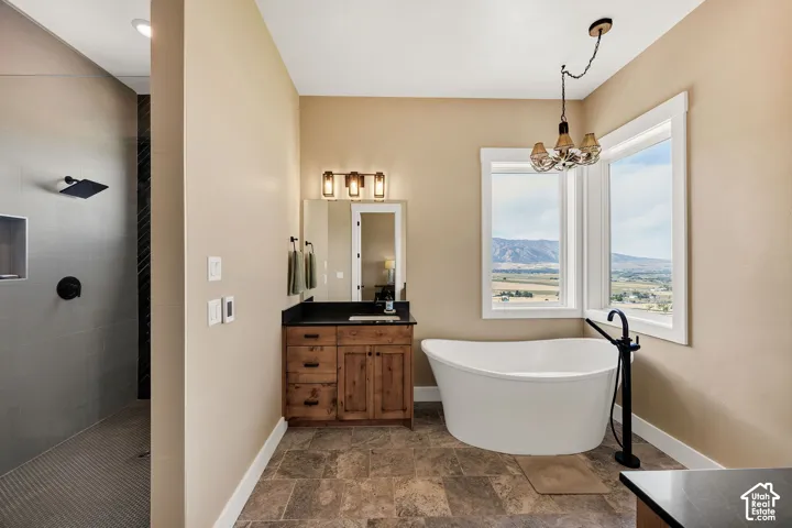 Freestanding soaking tub with a floor-mounted matte black faucet, walk-in shower with large-format tile, wood-finish vanity cabinetry with a stone countertop, and bay windows featuring mountain scenery