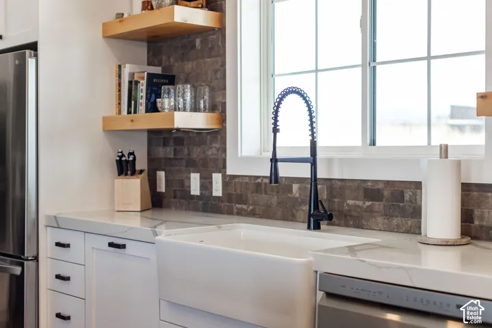 Kitchen featuring a white farmhouse sink, matte black gooseneck faucet, light-toned stone-finish countertops, textured stone backsplash, and natural wood-finish floating shelves