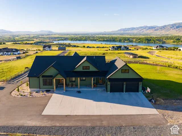 Multi-gabled residence with green vertical siding, dark roof shingles, attached three-car garage, covered front porch with timber posts, and expansive acreage featuring a mountain backdrop and water access