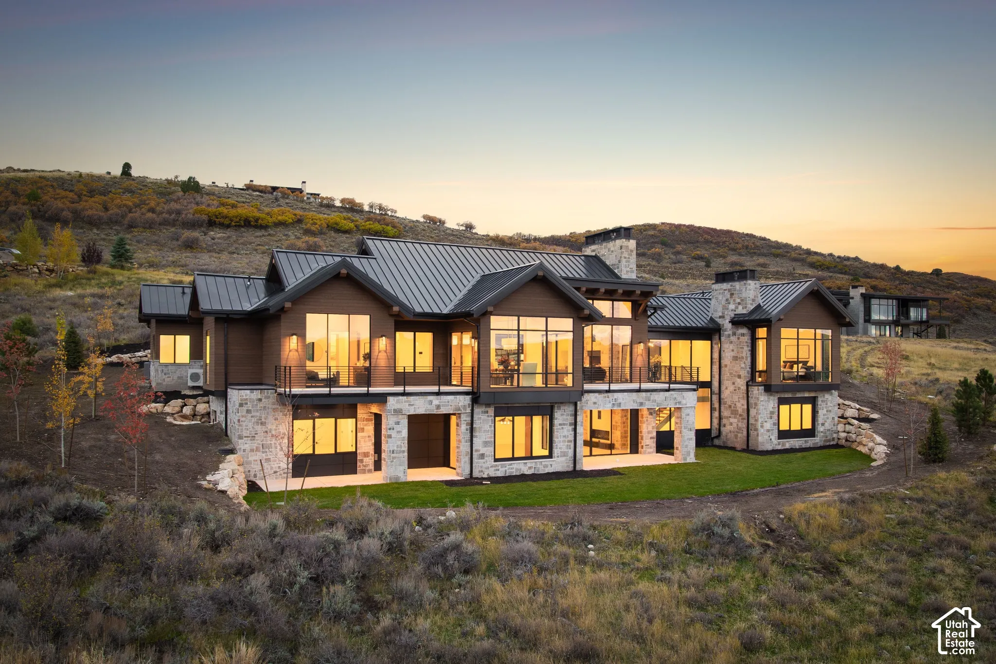 Custom-built exterior featuring stone masonry, metal roof, extensive floor-to-ceiling windows, wrap-around balcony with iron railings, and a covered lower-level patio