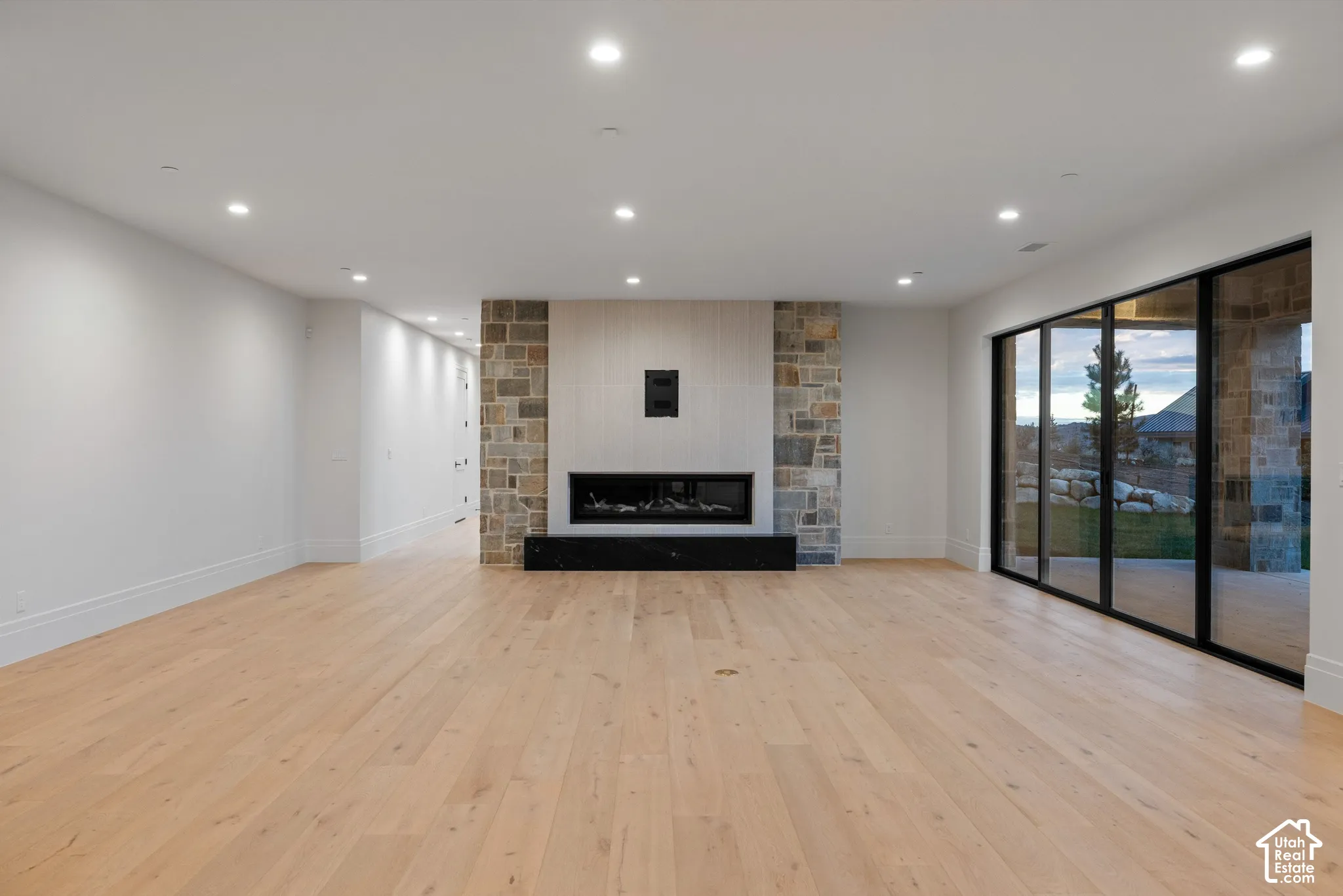 Open-concept interior featuring light wood-finish flooring, recessed lighting, and a floor-to-ceiling stone fireplace surround with a linear hearth