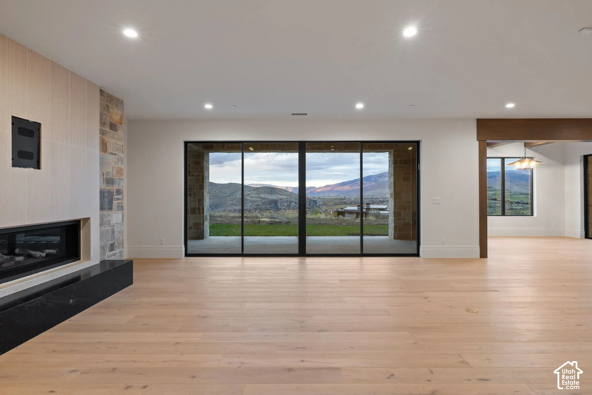 Open-concept interior featuring wood-finish flooring, recessed lighting, a stone-accented linear fireplace, and expansive multi-panel glass doors opening to a mountain backdrop