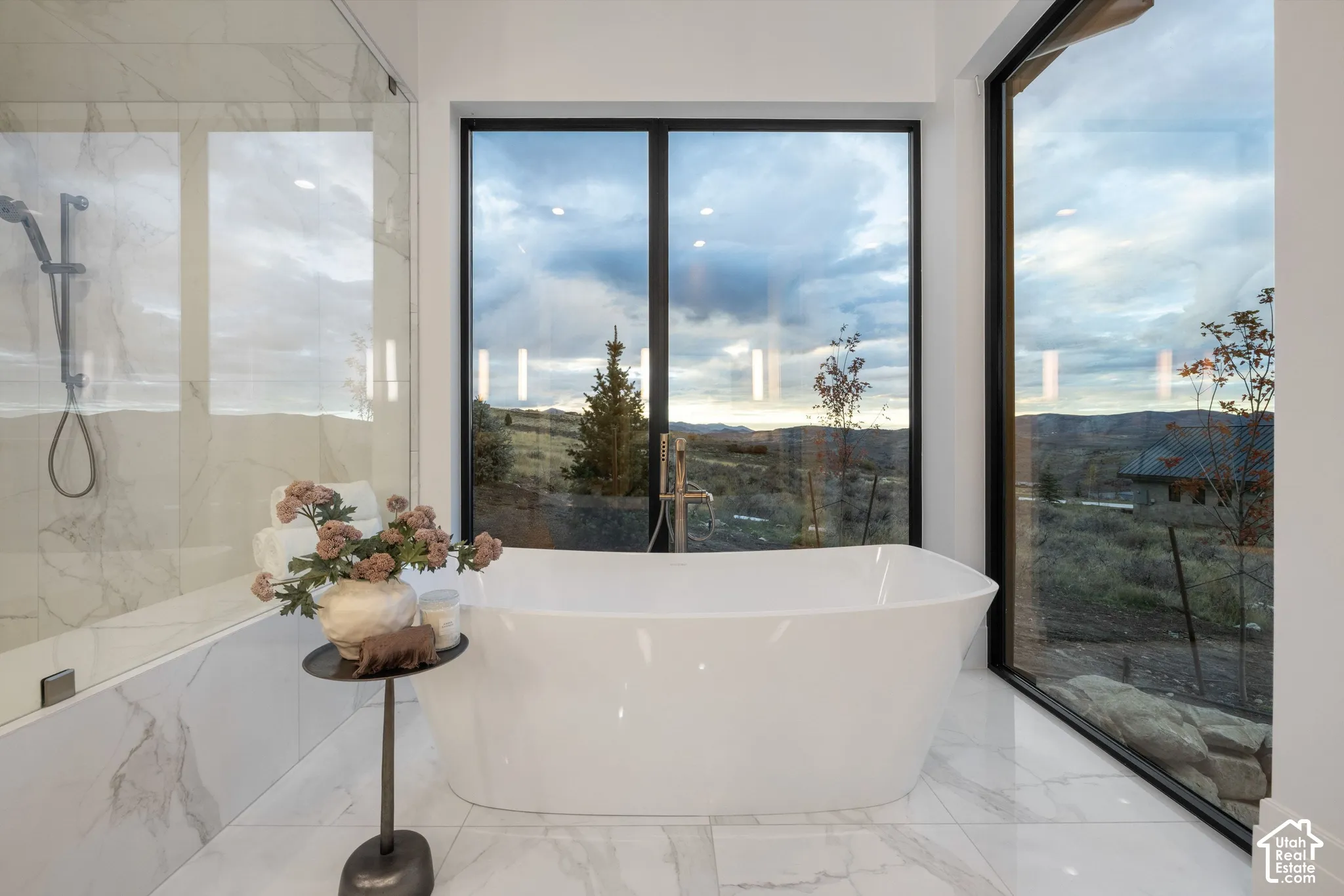 Freestanding soaking tub, large-format tile flooring, wall-to-wall glass shower enclosure with stone-finish surround, floor-to-ceiling windows with mountain scenery, and matte black fixtures