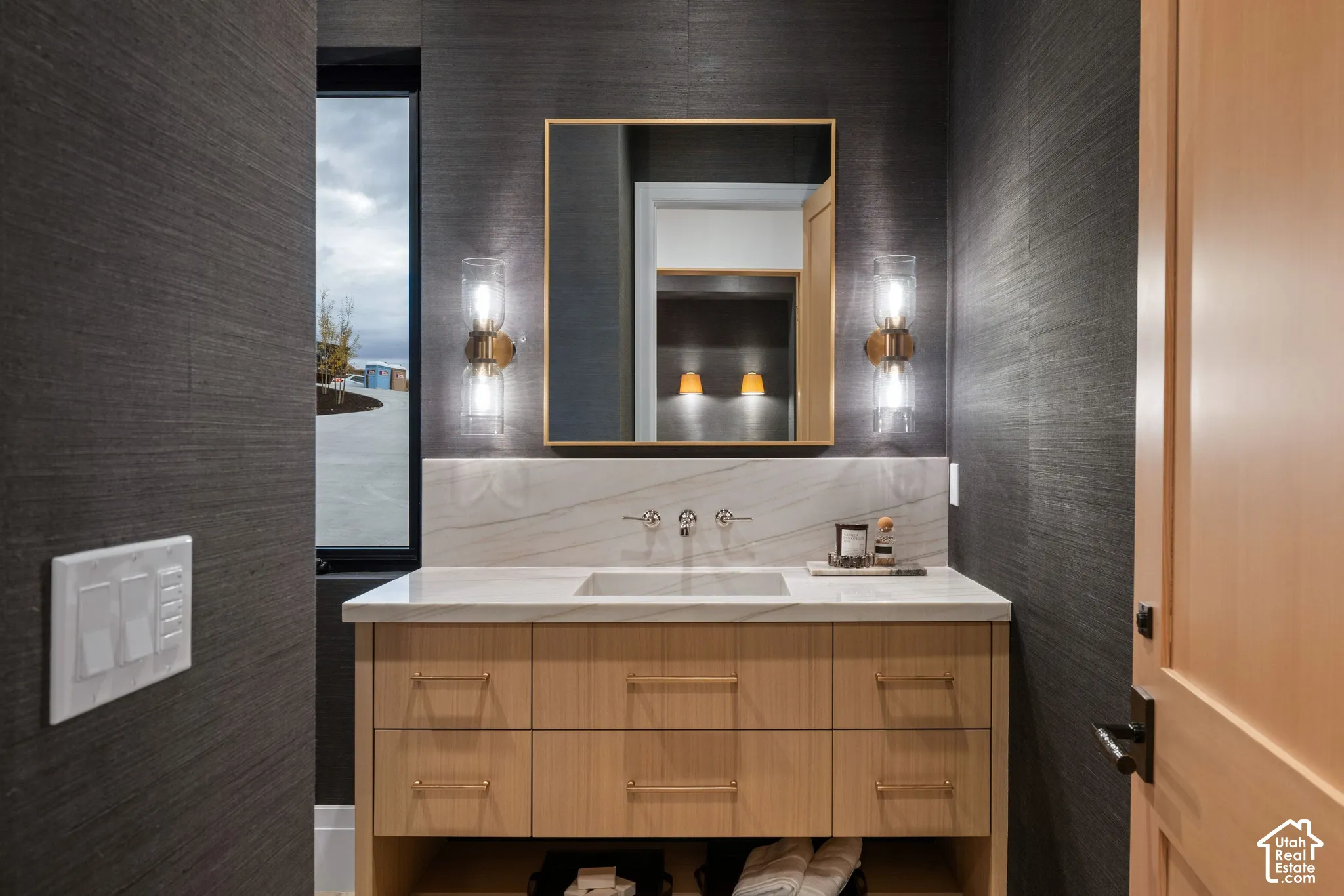 Custom floating wood-finish vanity with an integrated basin, stone-surface backsplash, textured dark wall coverings, and dual glass wall sconces