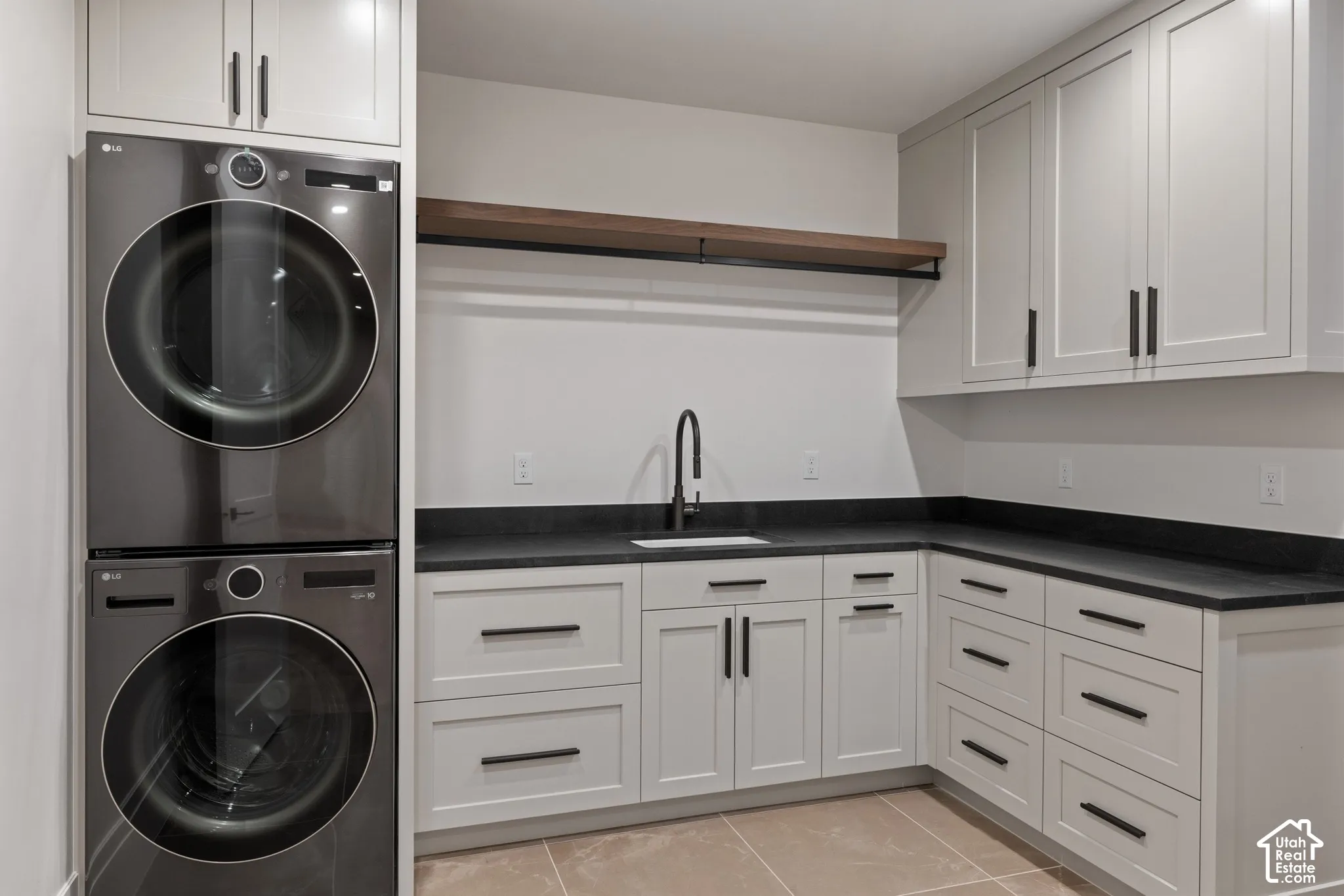 Laundry room featuring shaker-style cabinetry, matte black hardware, stone-finish countertops, an undermount sink with a high-arch faucet, and a wood-finish floating shelf
