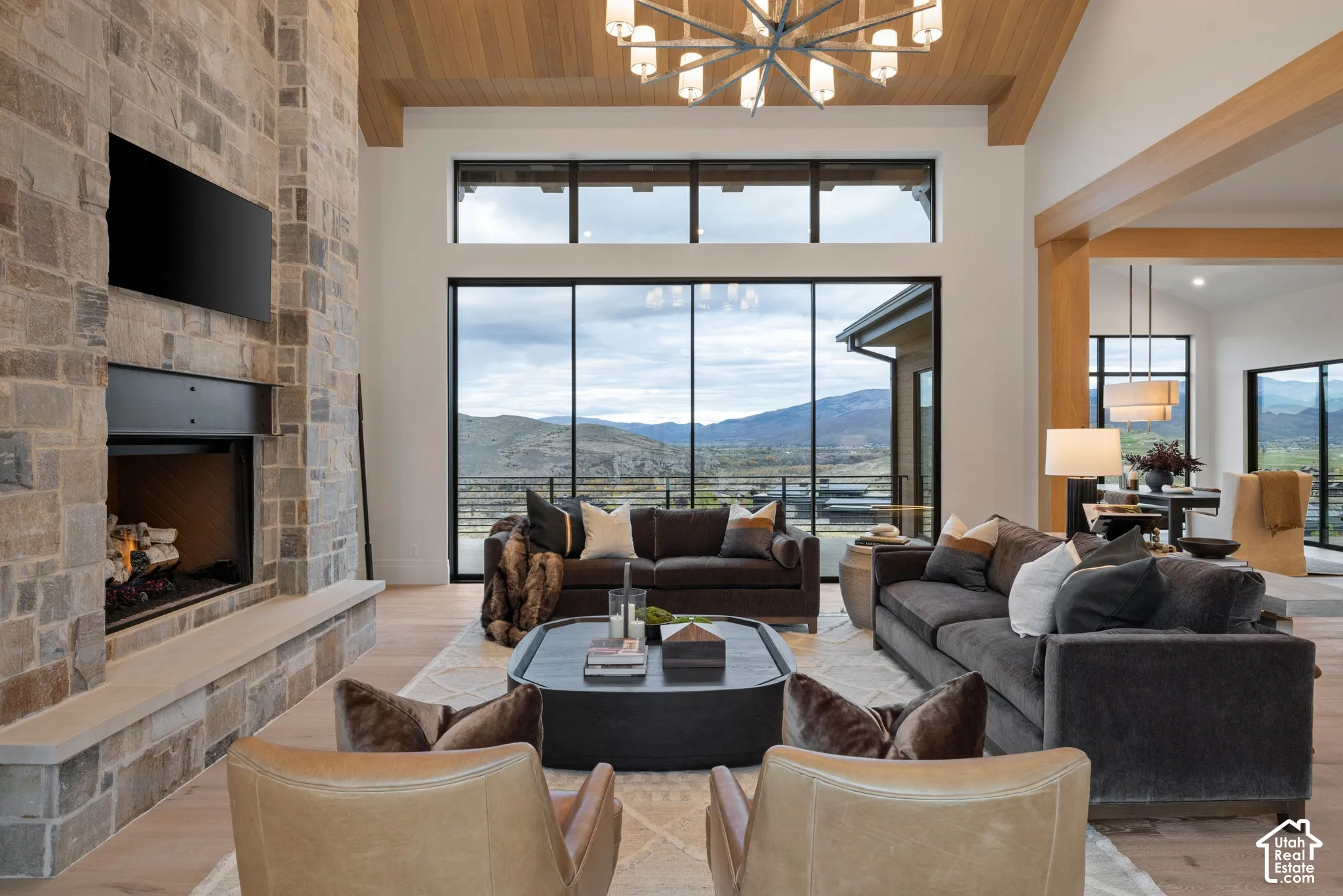 Floor-to-ceiling multi-panel windows, stone-clad fireplace with timber mantel, wood-plank ceiling beams, light-toned wood-finish flooring, and expansive mountain scenery