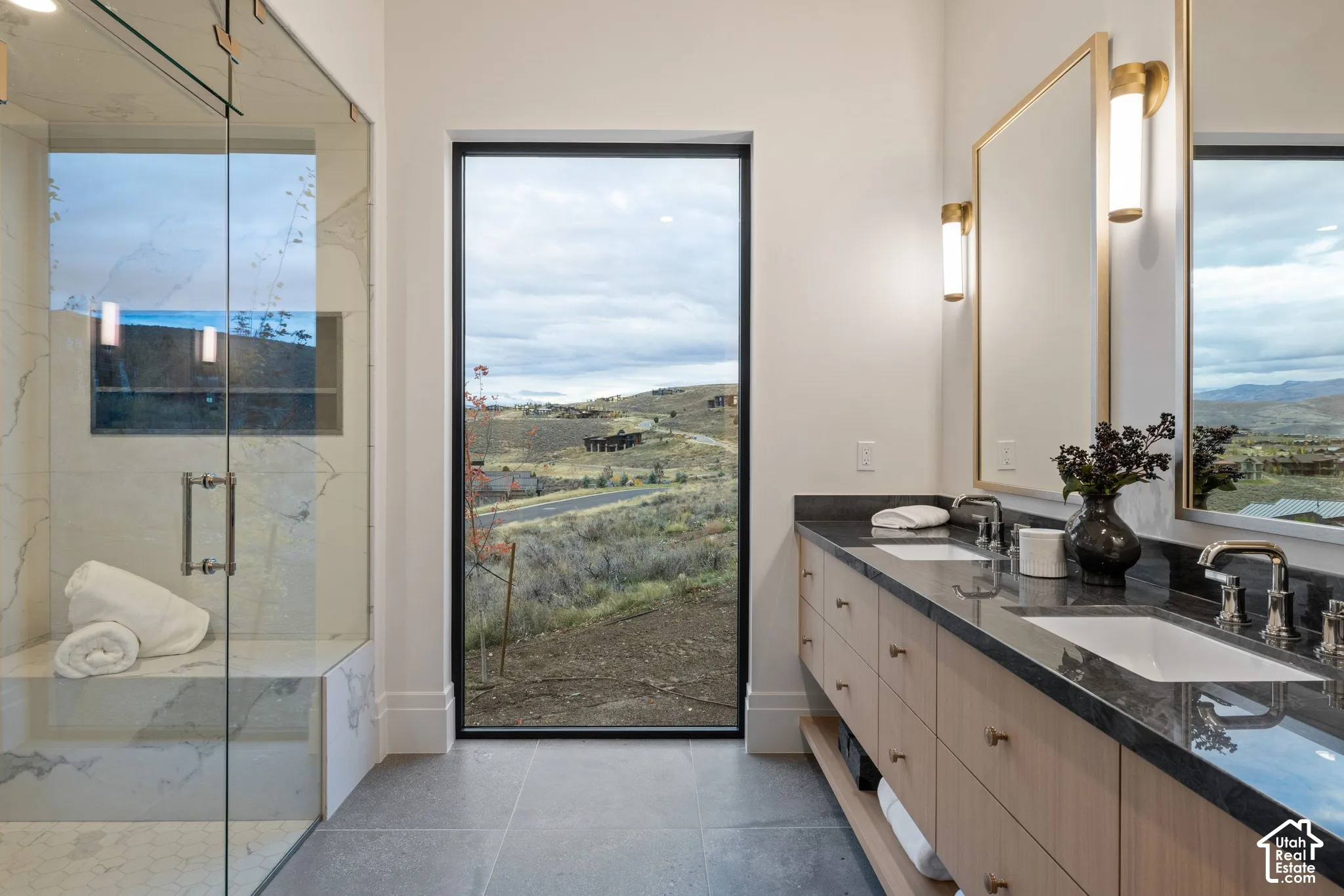 Floor-to-ceiling window, dual-sink vanity with dark stone countertop, light wood cabinetry, and a walk-in glass-enclosed shower with integrated bench seating