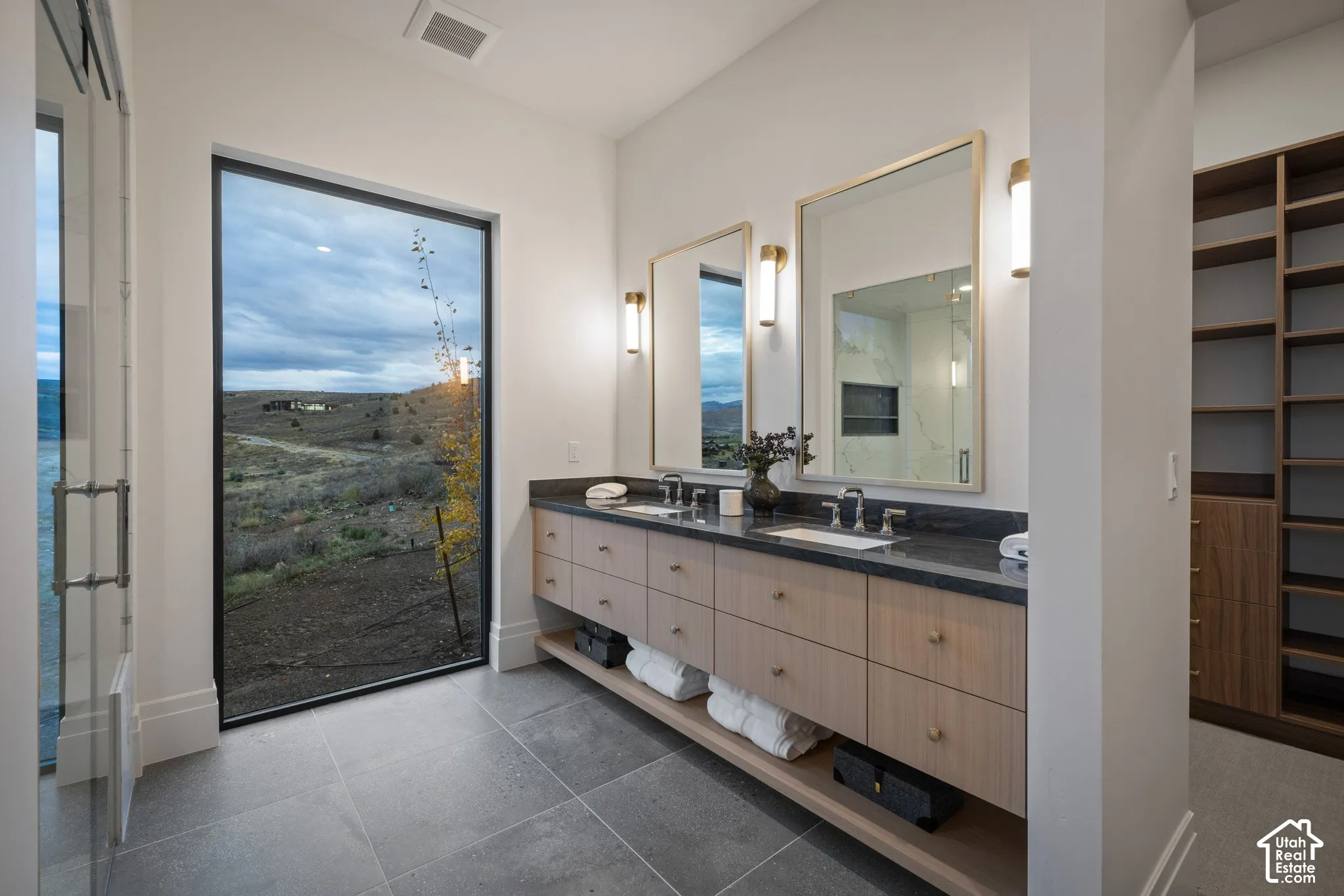 Floating double vanity featuring light-toned wood cabinetry, dark stone countertops, dual under-mount sinks, and wall-mounted metallic fixtures