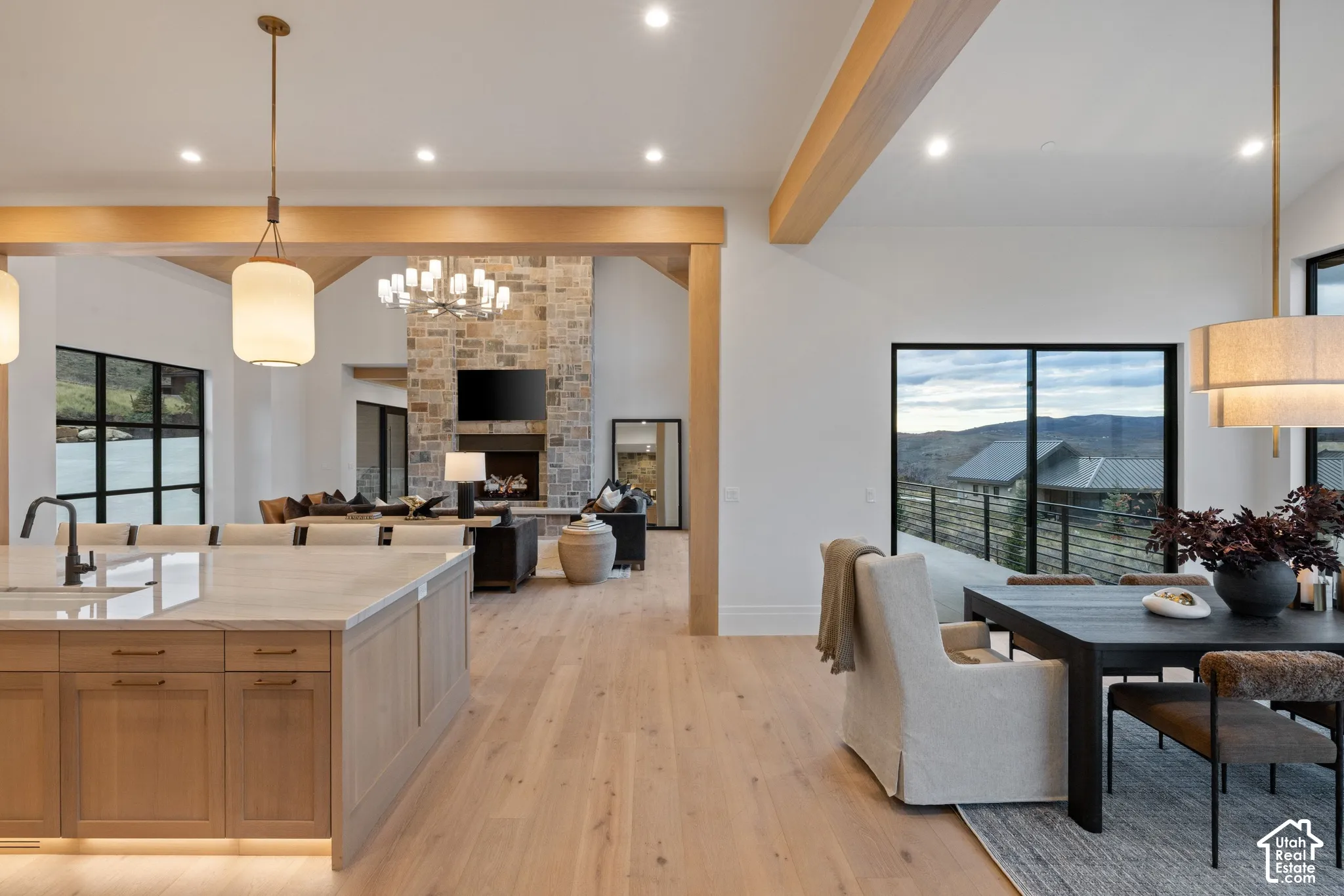 Open-concept living space featuring wood-finish flooring, exposed natural wood beams, stone-clad fireplace, kitchen island with stone countertop, and black-frame windows