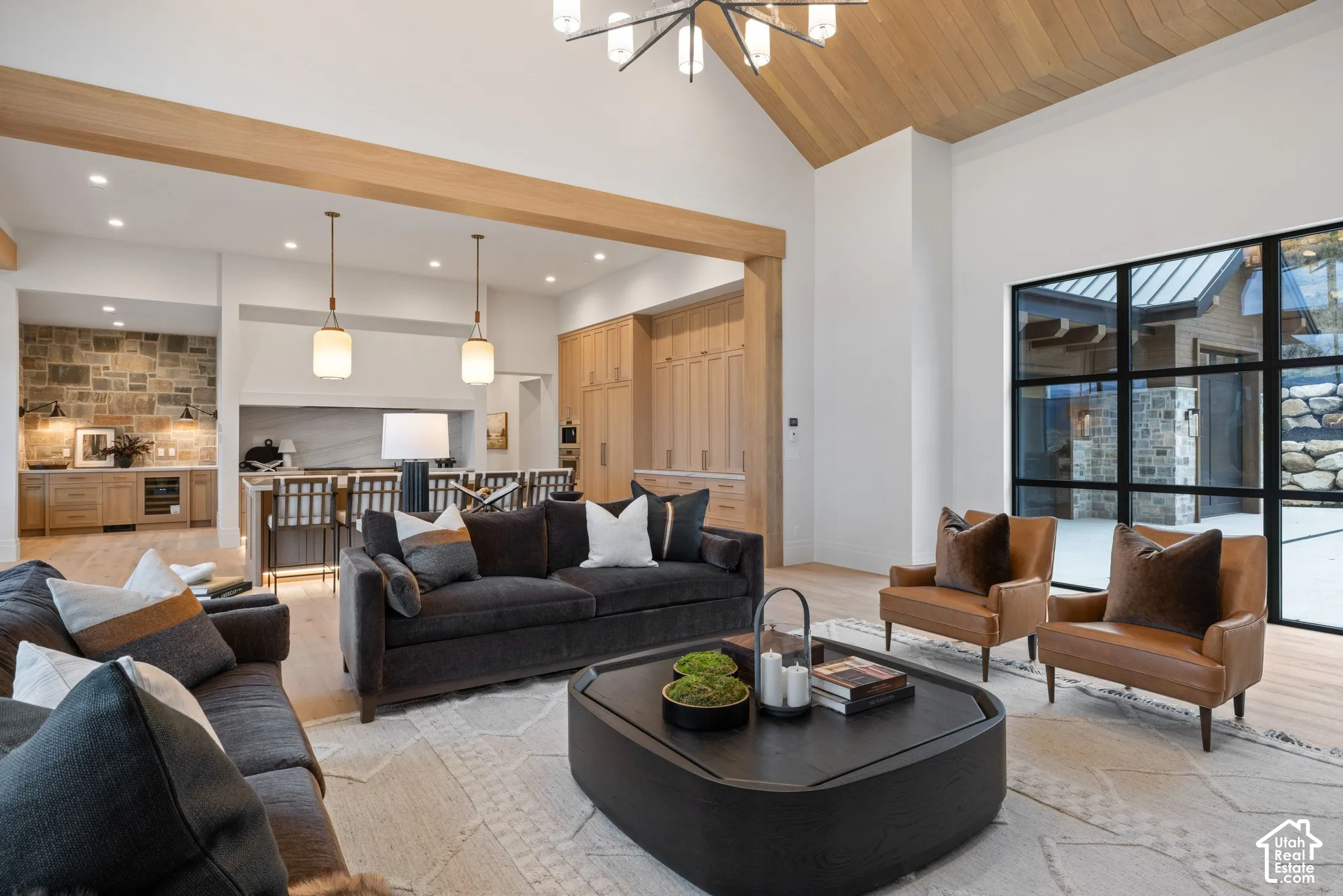 Vaulted ceiling with wood-paneled accents, exposed wood structural beam, floor-to-ceiling black-framed windows, stone-faced kitchen wall, and integrated wood-finish cabinetry