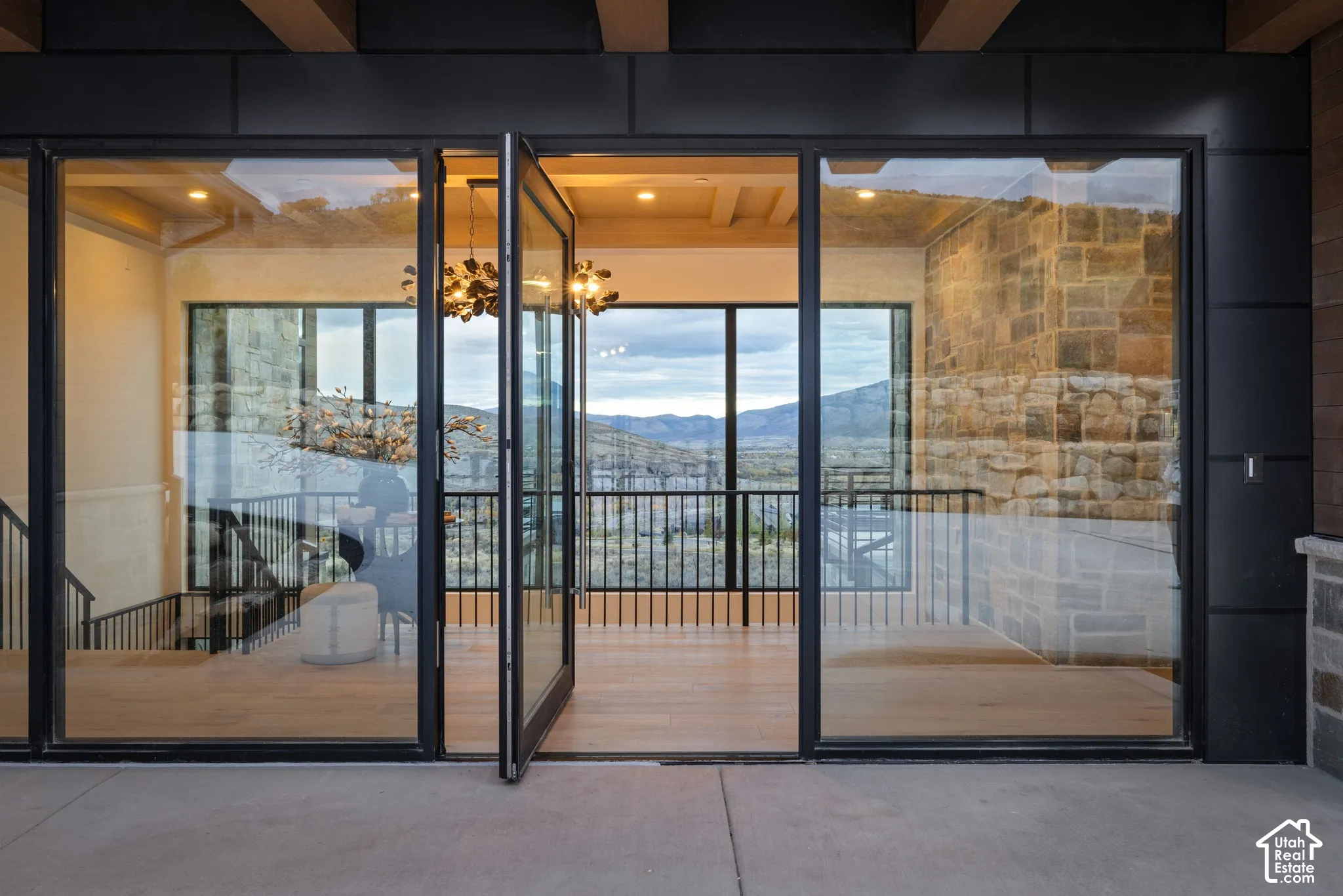 Floor-to-ceiling glass panel door system with black metal framing, exposed wood-beam ceiling detailing, stone veneer masonry, and wood-finish flooring