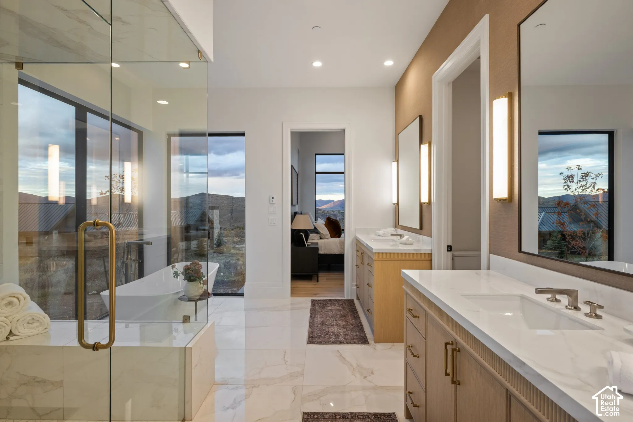 Large-format stone-pattern flooring, dual vanity cabinetry with stone countertops, glass-enclosed shower, freestanding soaking tub, and wall-mounted light fixtures