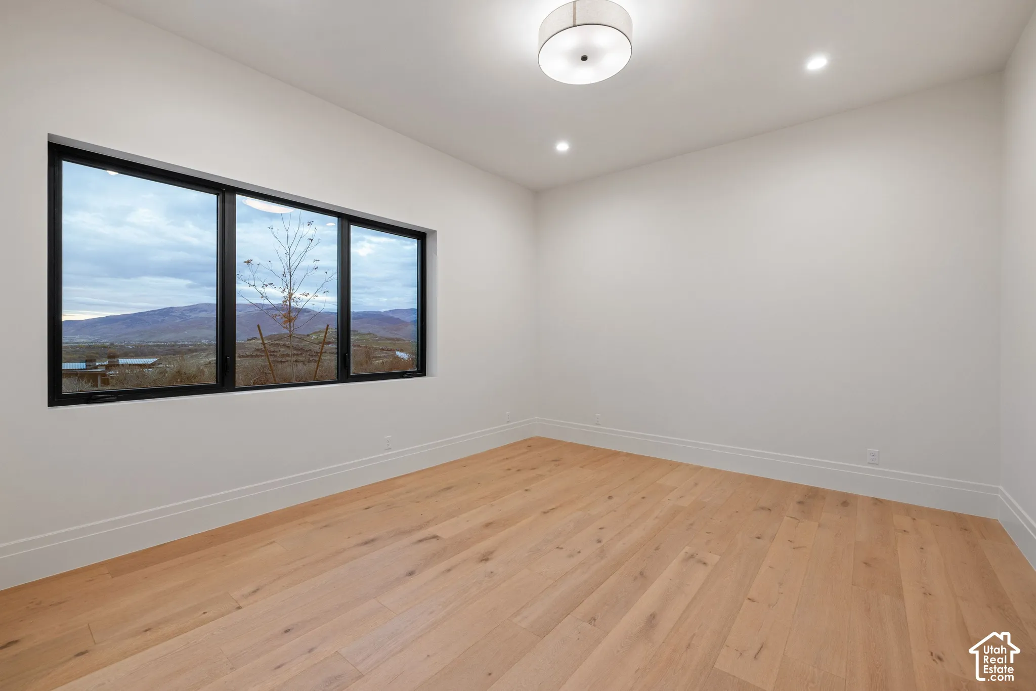 Neutral interior space featuring wood-finish flooring, recessed lighting, a drum-style ceiling fixture, and a large window with black framing showcasing mountain scenery