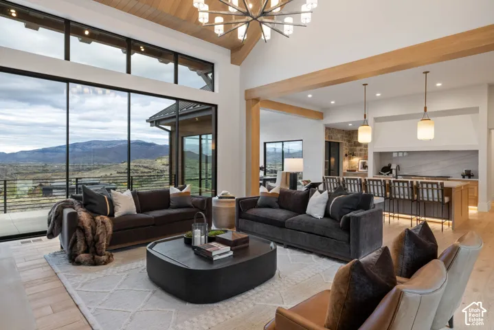 Vaulted ceilings with exposed timber beams, expansive floor-to-ceiling windows, wood-finish flooring, custom chandelier lighting, and a stone-accented kitchen backsplash