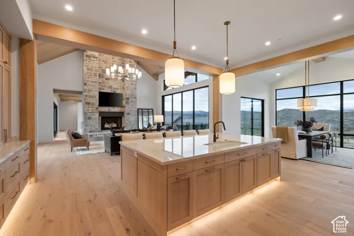 Open-concept kitchen featuring an oversized island with quartz countertops, custom wood-finish cabinetry, floor-to-ceiling windows, vaulted ceilings with wood beams, and a floor-to-ceiling stone fireplace