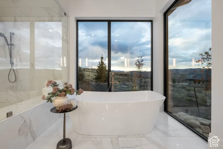 Freestanding soaking tub, large-format tile flooring, wall-to-wall glass shower enclosure with stone-finish surround, floor-to-ceiling windows with mountain scenery, and matte black fixtures