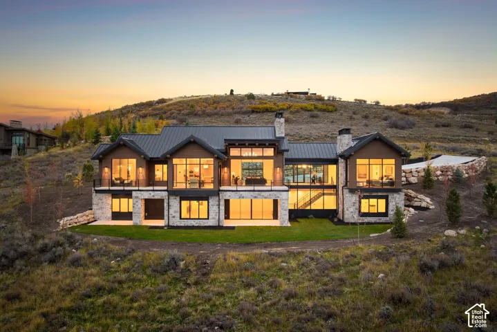 Custom residence featuring a multi-gabled metal roof, stone masonry siding, expansive floor-to-ceiling windows, integrated balcony with glass railing, and structural timber detailing