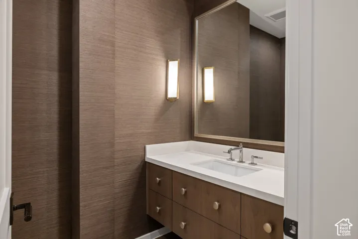 Textured wall covering featuring a wood-finish floating vanity, integrated rectangular sink, solid surface countertop, and metal wall-mounted lighting fixtures
