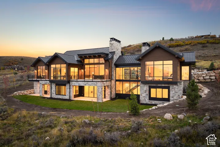 Multi-level mountain residence featuring stone veneer siding, floor-to-ceiling windows, metal roofing, elevated balcony with glass railing, and dual exterior stone chimneys
