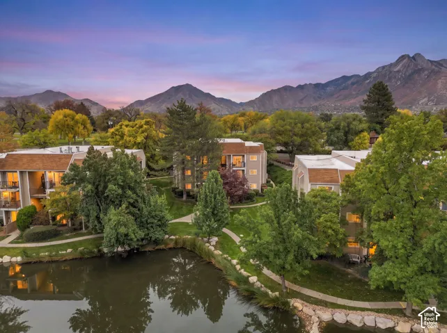 Multi-level residential structures featuring private balconies, situated on a landscaped lot with a pond, paved pathways, and prominent mountain backdrop