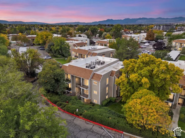 Multi-story building featuring shingled rooflines, exterior balconies, mature deciduous trees, and mountain scenery